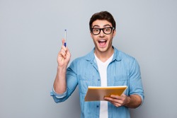 Handsome, attractive, glad, positive, funny guy in glasses with wide open mouth finally found a solution how to make exercise, having raised pen and notebook in hands, isolated on grey background