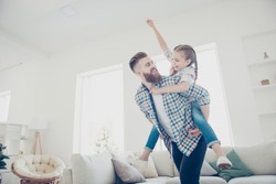 Cheerful father carrying on back little playful joyful kid with raised fist celebrating victory in game, family with one parent spending time in modern house with interior, babysitting concept