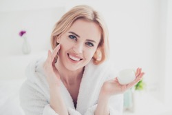 Closeup portrait of adult beautiful attractive blonde cute mature smiling woman wearing white bathrobe spreading face lotion cream gel on her face. Keeping and showing cream jar in other hand