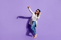 Full size body of happy smiling girl in jeans overcoat street look shooting self portrait on front camera of smart phone posing isolated on violet background