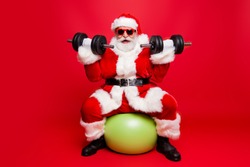 Cheerful sporty muscular virile strong Santa in white fluffy gloves fur coat sitting on pilates ball work out ready for sale promo discount wishing merry x mas isolated on red background fun joy