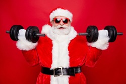 Portrait of nice optimistic positive modern stylish strong sporty muscular virile Saint Nicholas in gloves fur white red winter clothes holding lifting two big dumbbells isolated over red background