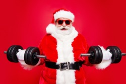 Portrait of stylish virile strong muscular sporty Saint Nicholas in eyeglasses gloves fur white red winter coat clothes holding lifting two big dumbbells in arms striving isolated on red background