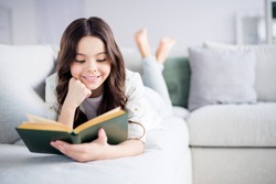 Portrait of her she nice attractive lovely charming cute cheerful cheery focused wavy-haired preteen girl lying on divan reading novel in light white interior living-room indoors