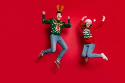 Full length photo of lady and guy jumping excited by x-mas discounts wear ugly ornament jumpers and headwear isolated red color background