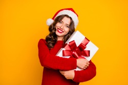 Pretty overjoyed lady holding large giftbox close to chest wear knitted jumper and santa hat isolated yellow background