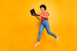 Full length photo of excited funny funky curly hair girl use her laptop find news information about sales jump wear denim jeans stylish trendy sneakers t-shirt isolated over yellow color background