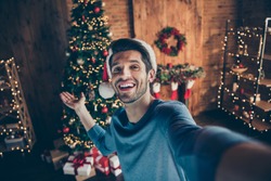 Close up photo of happy joy guy wear santa claus hat make selfie hold hand invite to his x-mas party celebration in house with christmas decoration illuminations tinsels wreath