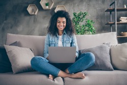 Photo of pretty amazed dark skin curly lady notebook on knees crossed legs texting friends read good news sitting comfy couch casual denim outfit living room indoors