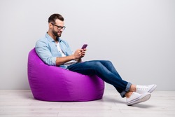 Full length profile photo of positive guy sitting comfy violet armchair holding telephone chatting friends wear specs casual denim outfit isolated grey color background