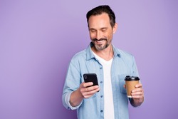 Portrait of cheerful positive man have free time relax after work use smartphone check social network hold mug with hot beverage wear denim jeans shirt isolated over violet color background