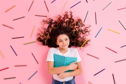 Top view above high angle flat lay flatlay lie concept portrait of nice beautiful small little wavy-haired girl hugging book many colorful pencils isolated over pink pastel color background