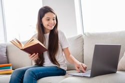 Photo of little pretty pupil school lady turn on skype laptop notebook online lesson hold open book ready answer teacher questions sit sofa distance quarantine study comfort room indoors