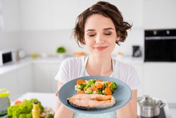 Close up photo of pretty housewife lady chef hold ready grilled salmon trout fillet steak with garnish cook dinner one person portion eyes closed wear apron t-shirt modern kitchen indoors