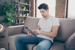 Portrait of his he nice attractive focused brunette guy sitting on divan reading ebook using app web service at modern industrial loft brick interior style living-room