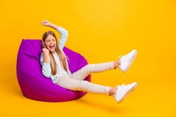 Full size profile photo of beautiful little lady good mood sit cozy violet bean bag rejoicing win raise fists summer holidays wear casual shirt pants shoes isolated yellow color background