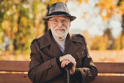 Photo of cheerful retired old grey haired grandpa street central park sit bench positive emotions enjoy sunny day weather hold walking cane stick wear autumn glasses jacket hat outside