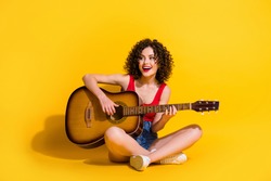 Portrait photo of hipster female musician with curly hair singing song holding keeping playing melody guitar chords smiling sitting down isolated on vivid yellow color background