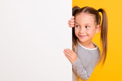Portrait of young happy smiling curious little girl child kid behind white wall banner look in copyspace isolated on yellow color background
