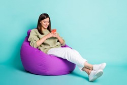 Photo portrait full body view of smiling girl chatting holding phone in two hands sitting in beanbag chair isolated on vivid turquoise colored background