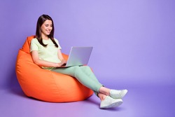 Photo portrait full body view of woman with laptop sitting in orange beanbag isolated on vivid purple colored background