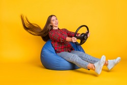 Portrait of beautiful trendy cheerful girl sitting in bag chair holding steering wheel wind blow hair isolated on bright yellow color background