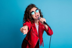 Photo of crazy carefree lady open mouth sing hold mic direct finger on you wear round sunglass isolated on blue color background
