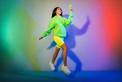 Full length body size view of attractive cheerful girl jumping using device isolated over multicolor vivid neon light background
