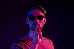 Portrait of attractive mysterious guy showing shh sign keep silence isolated over dark neon violet color background