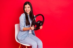 Portrait of pretty cheerful girl sitting in chair holding in hands steering wheel study course isolated over bright red color background