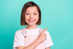 Photo of adorable pretty schoolgirl dressed pink overall pointing finger empty space smiling isolated turquoise color background