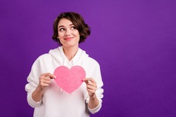 Photo of pretty sweet young woman dressed white pullover holding pink heart looking empty space smiling isolated violet color background