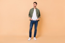 Full size photo of dreamy young man look empty space hold hands pockets jeans isolated on beige color background