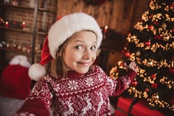 Photo of cute cheerful blogger small child girl shoot selfie demonstrate tree decor wear pullover santa hat indoors
