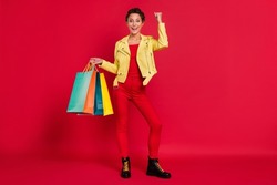 Photo of sweet lucky young woman dressed leather jacket holding shopping bargains rising fist smiling isolated red color background