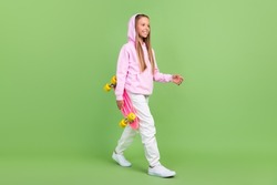Full length profile photo of nice little blond girl hold skate go wear hoodie trousers sneakers isolated on green background