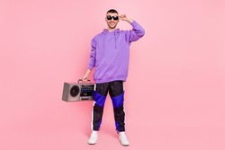 Full size photo of cool young brunet guy with boom box wear eyewear hoodie pants sneakers isolated on pink background