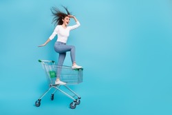 Photo of funky adorable woman wear white shirt riding shopping tray looking far away empty space isolated blue color background