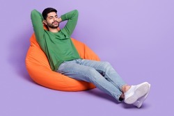 Full length photo of young latin man laying beanbag daydreaming chilling dressed stylish green garment isolated on purple color background