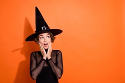 Photo of cute young girl astonished touch cheeks look empty space dressed trendy black halloween witch outfit isolated on orange background