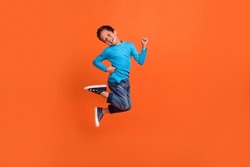 Full size photo of charming small pupil boy jump play raise fists celebrate dressed stylish blue look isolated on orange color background