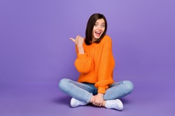 Full size photo of lovely young woman sit floor point copyspace dressed stylish orange knitted look isolated on purple color background