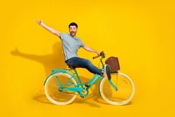 Side profile photo of funky crazy excited man wear gray t-shirt denim jeans sitting new retro bicycle raise palm hello friends isolated on yellow color background