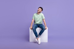 Full size photo of handsome young man sitting white podium look empty space dressed stylish gray outfit isolated on purple color background