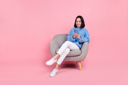 Full length photo of positive nice girl blue sweater white pants sitting on armchair look at smartphone isolated on pink color background