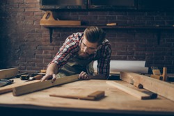 Bearded handsome cabinetmaker at the tabletop with tools.  Stylish craftsman with brutal hairstyle and saved glasses holding woodenplank at his workstation.
