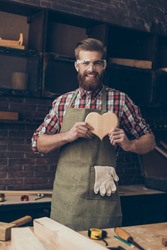 Happy handsome joiner in love holding and showing wooden heart near tabletop with tools.  Stylish young entrepreneur with beautyful hairstyle and saved glasses smile at his workstation. He love  job.