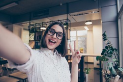 Portrait of excited cheerful smiling young pretty woman in spectacles making selfie photo and showing v-sign with two fingers