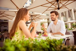 Young happy couple is sitting in light cafe`s terrace outdoors on a sunny day and enjoy, smiling, chatting with coffee and desserts