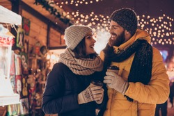 Having fun together at a christmas fairy. Young cheerful couple is having a walk with hot drinks, enjoying, dressed warm, looking at each other and laugh